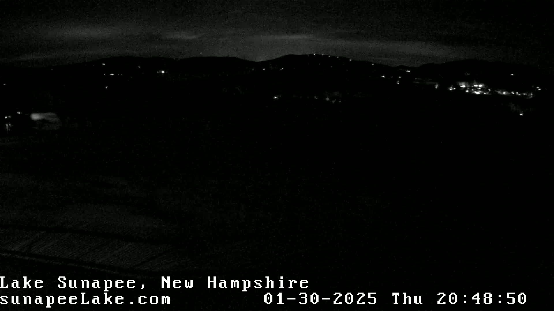 A nighttime view of a dark landscape with a few distant lights, under a mostly clear sky.