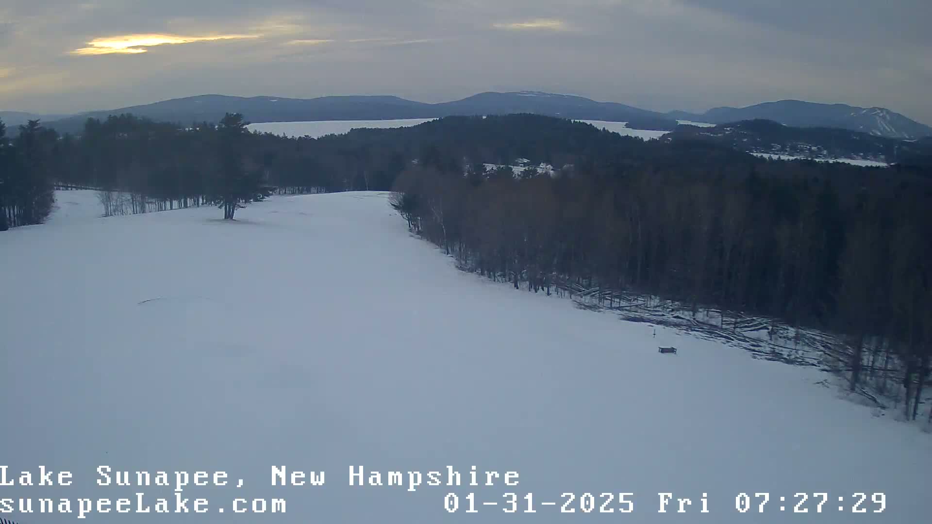 A snow-covered landscape with a lake and distant mountains under a cloudy, dusky sky.