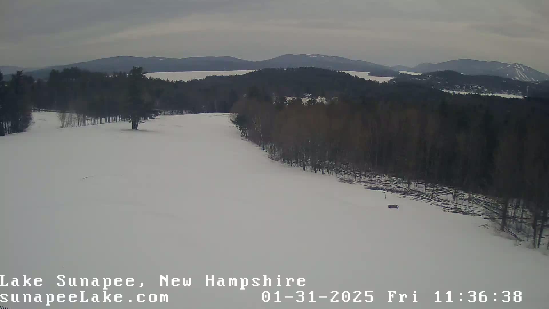 A snow-covered landscape with a distant view of a lake and mountains under an overcast sky.