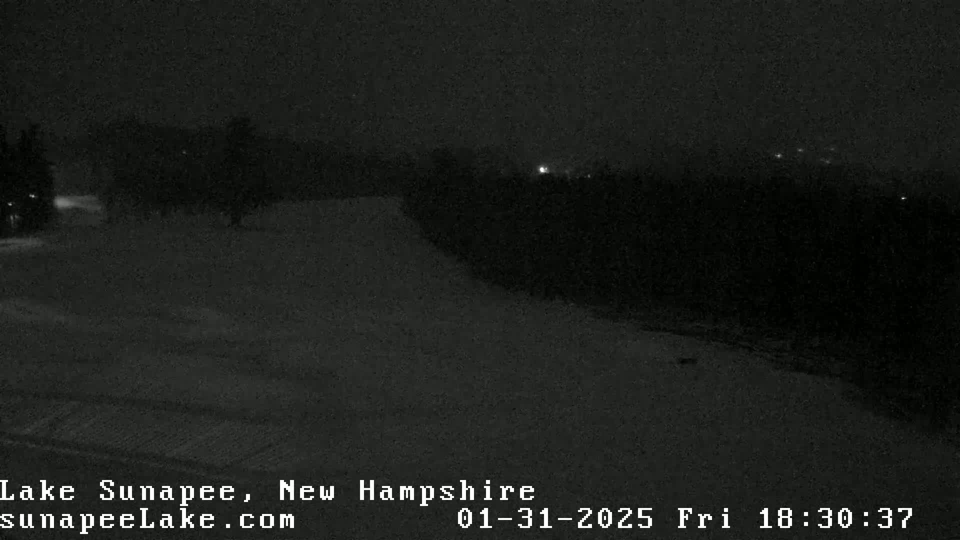 A nighttime, snowy scene shows a dark landscape with a few distant lights and some trees under a dark sky.