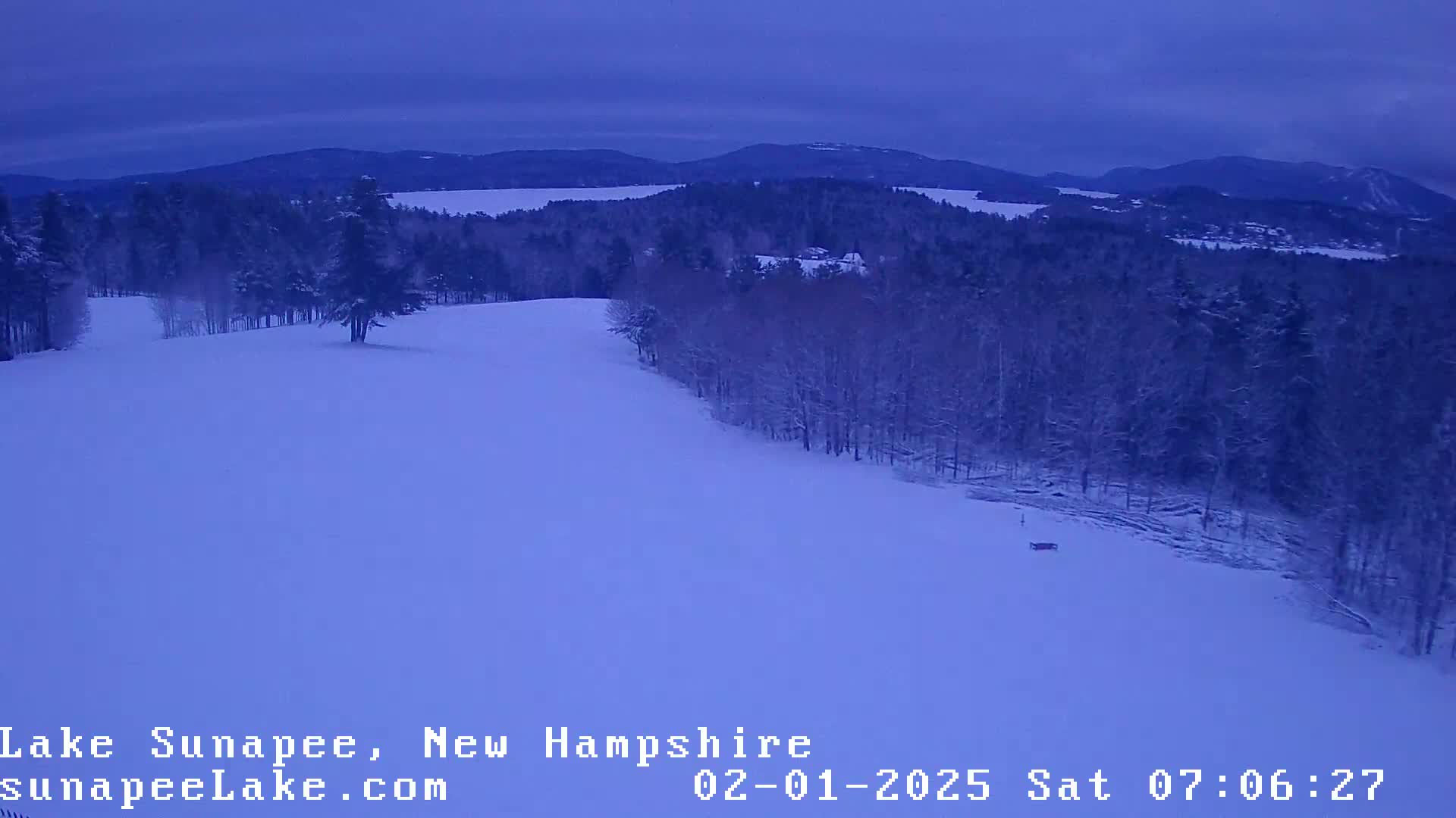 A snow-covered landscape at twilight shows a gently sloping hill lined with snow-covered trees leading to a distant mountain range.