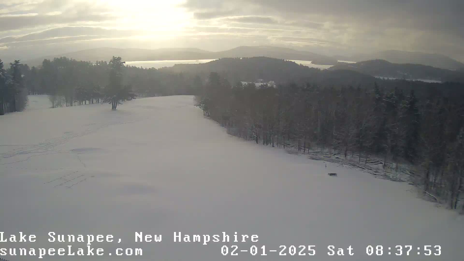 A snow-covered hill overlooks a lake and distant mountains under a partly cloudy sky.