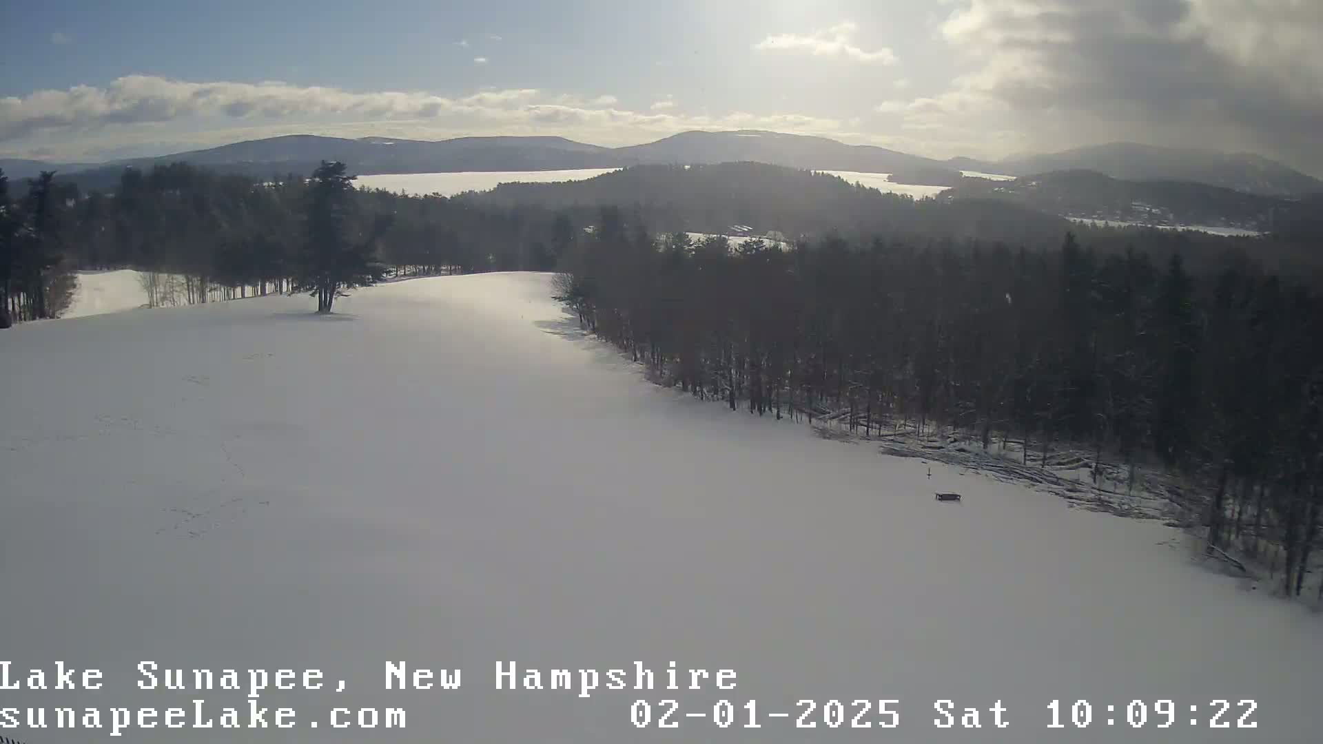 A snow-covered landscape with a line of trees and a body of water visible in the distance under a partly cloudy sky.