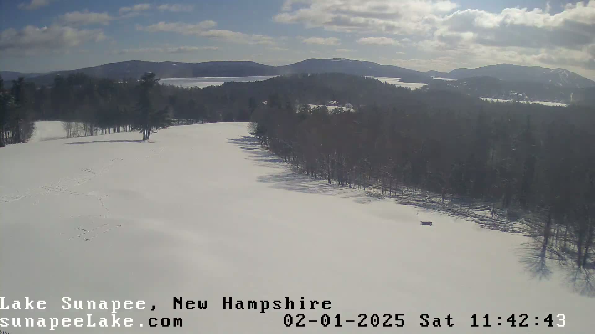 A snowy landscape with a partly sunny sky shows a view of a snow-covered hill leading down to a lake and distant mountains.