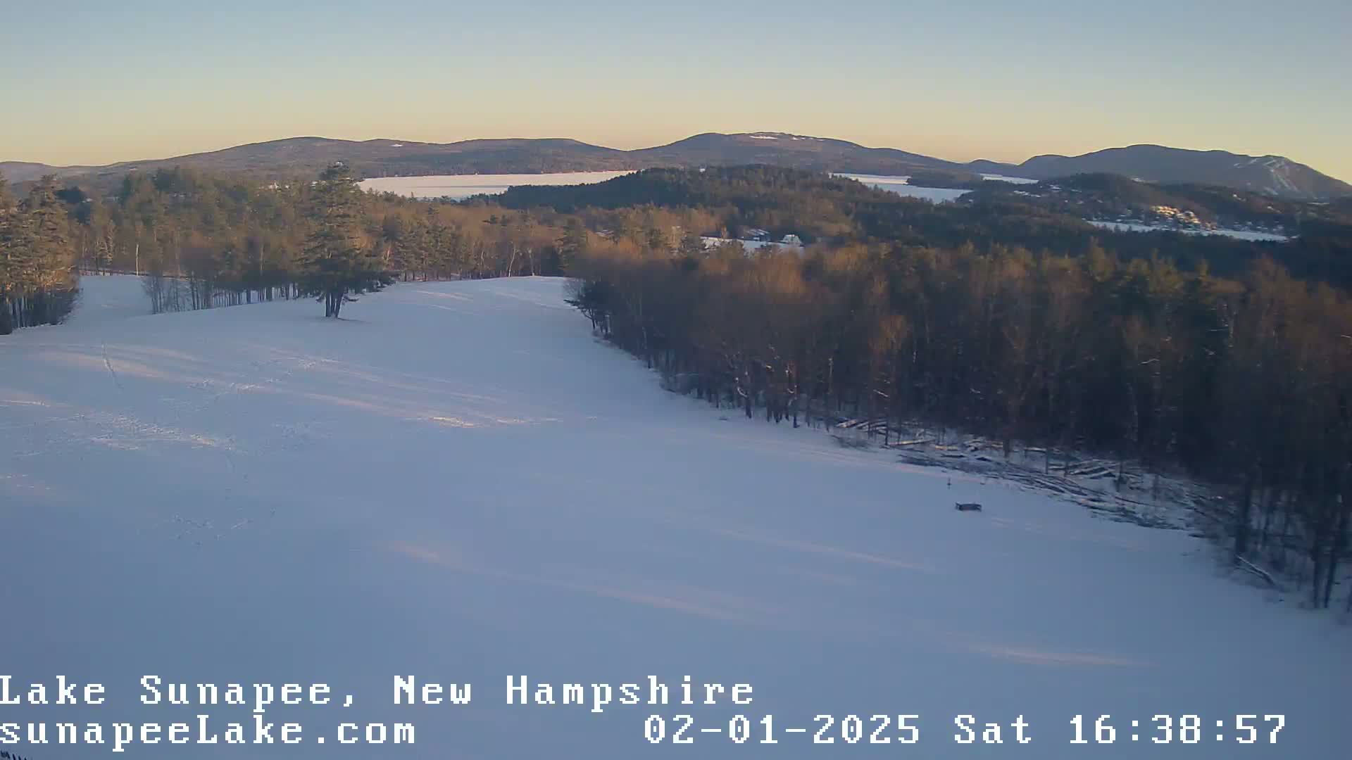 A snow-covered hillside slopes down toward a frozen lake, surrounded by dark evergreen trees under a clear, twilight sky.