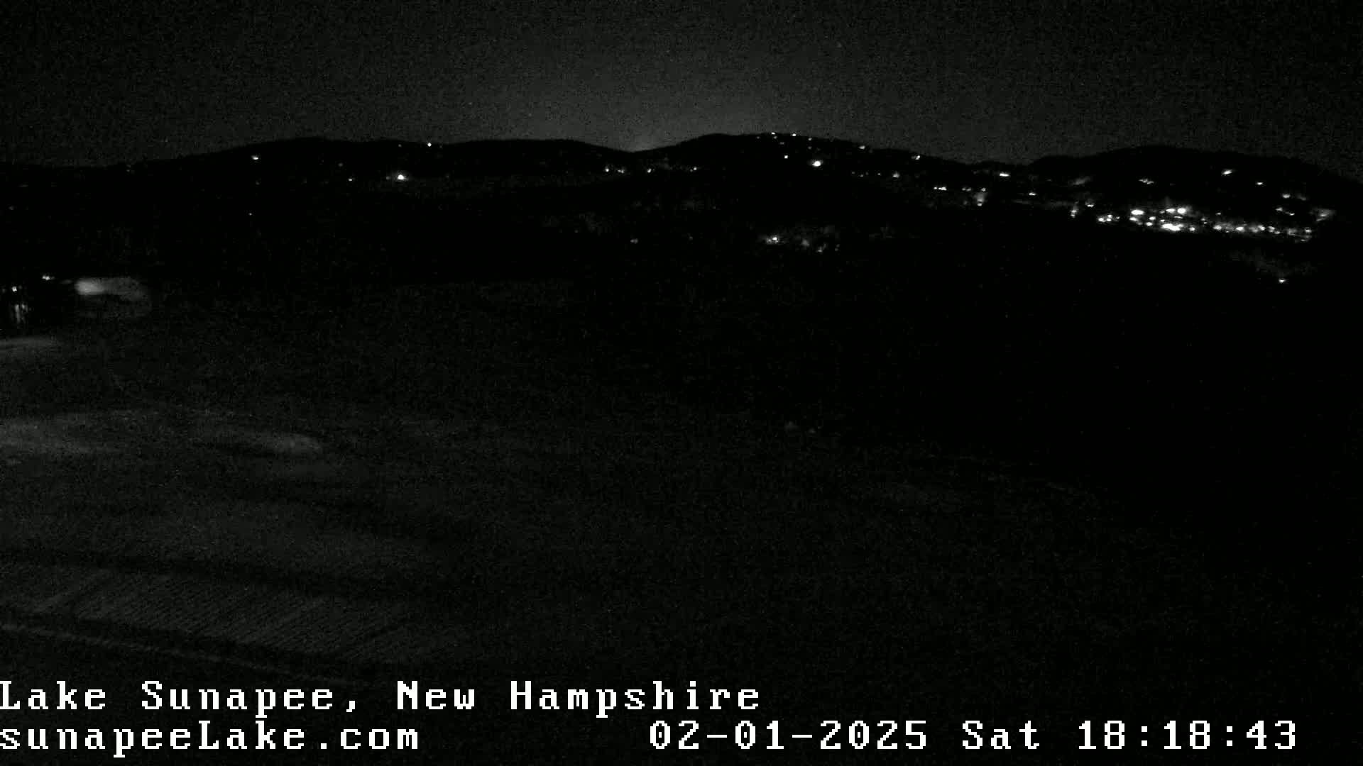 A nighttime, low-light view of a hilly landscape with scattered lights, possibly a small town, in the distance.