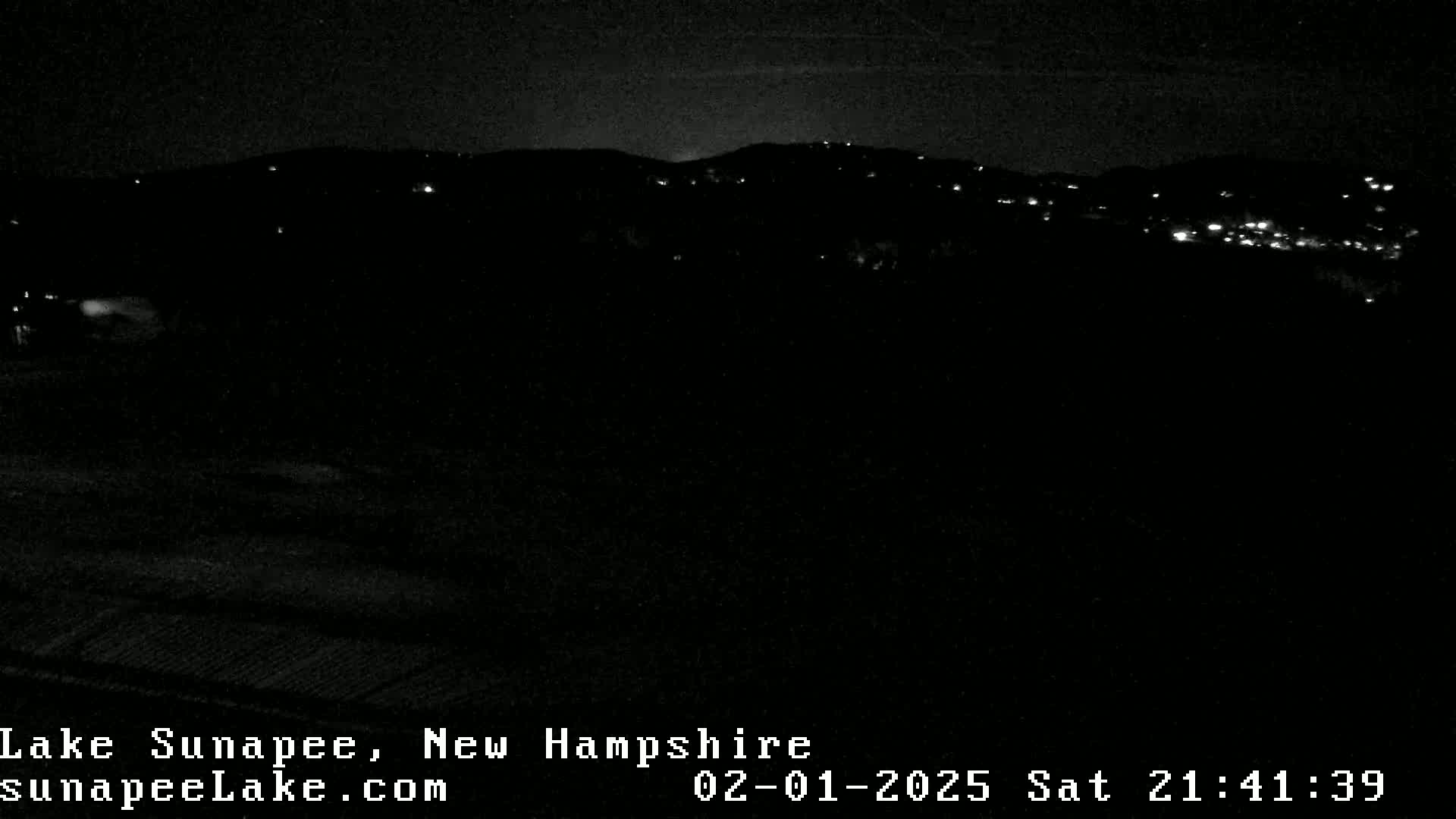 A nighttime view of sparsely lit hills and a dark foreground under a clear sky.