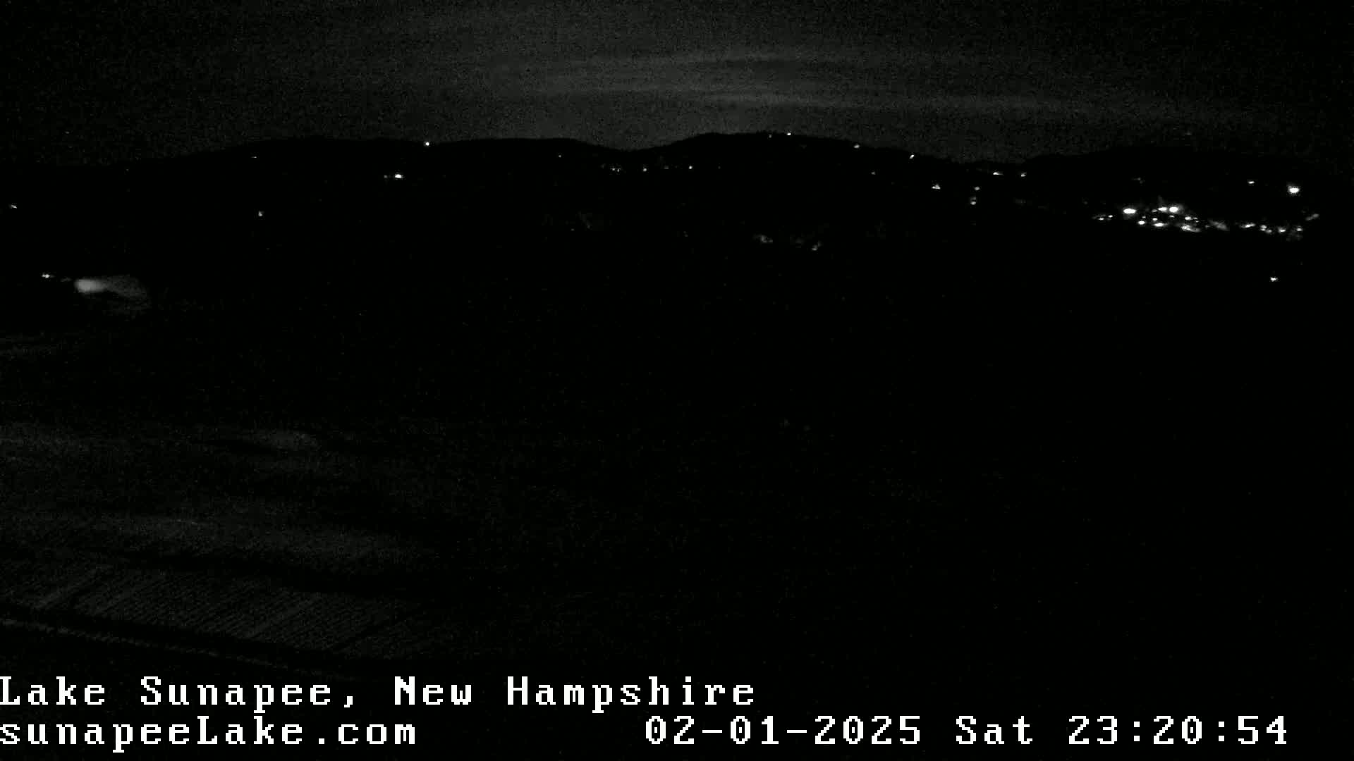 A nighttime, low-light view of a dark landscape with sparsely scattered lights in the distance under a mostly clear sky.