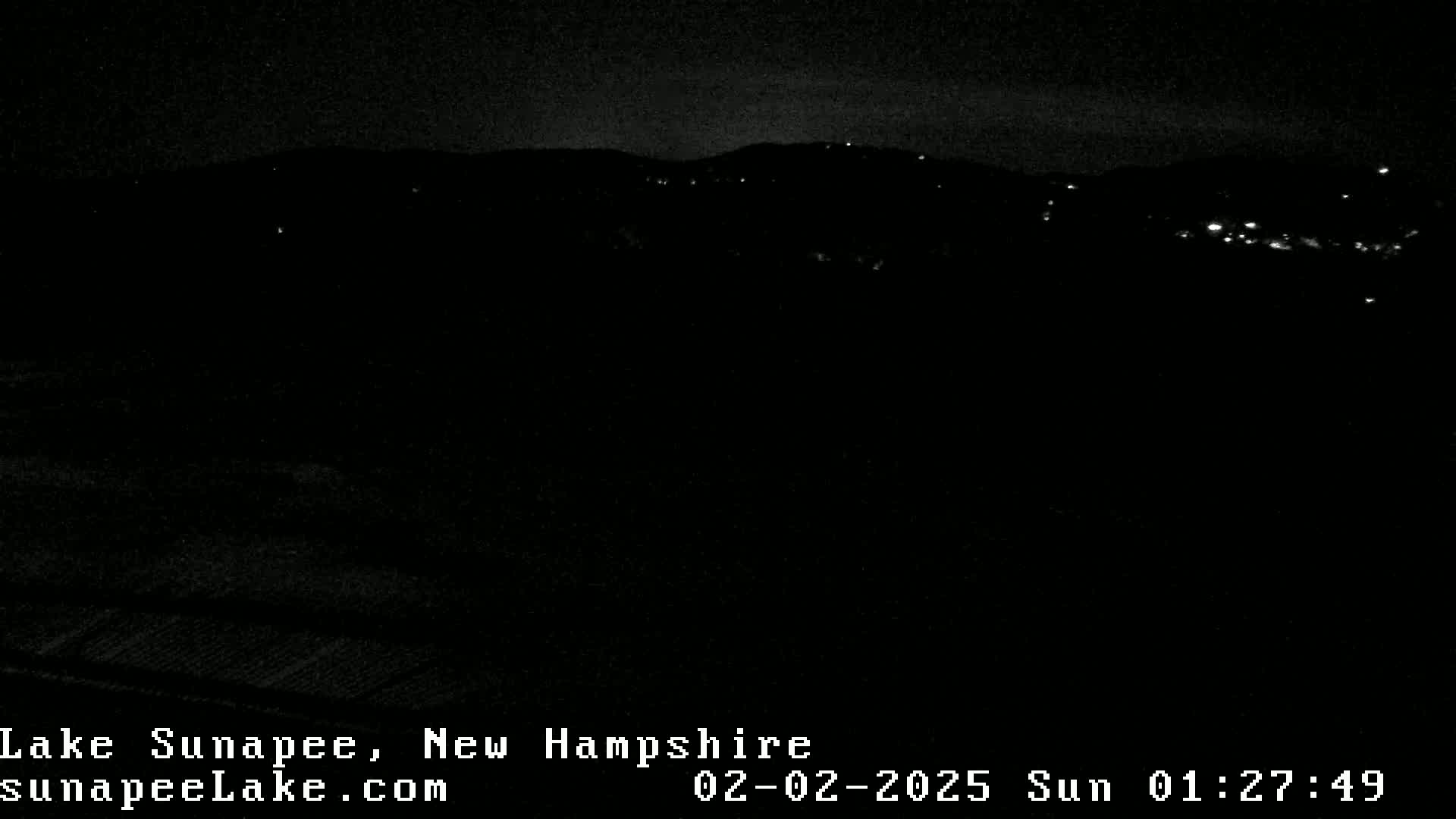 A nighttime view of a dark landscape with a few distant lights visible under a mostly clear sky.