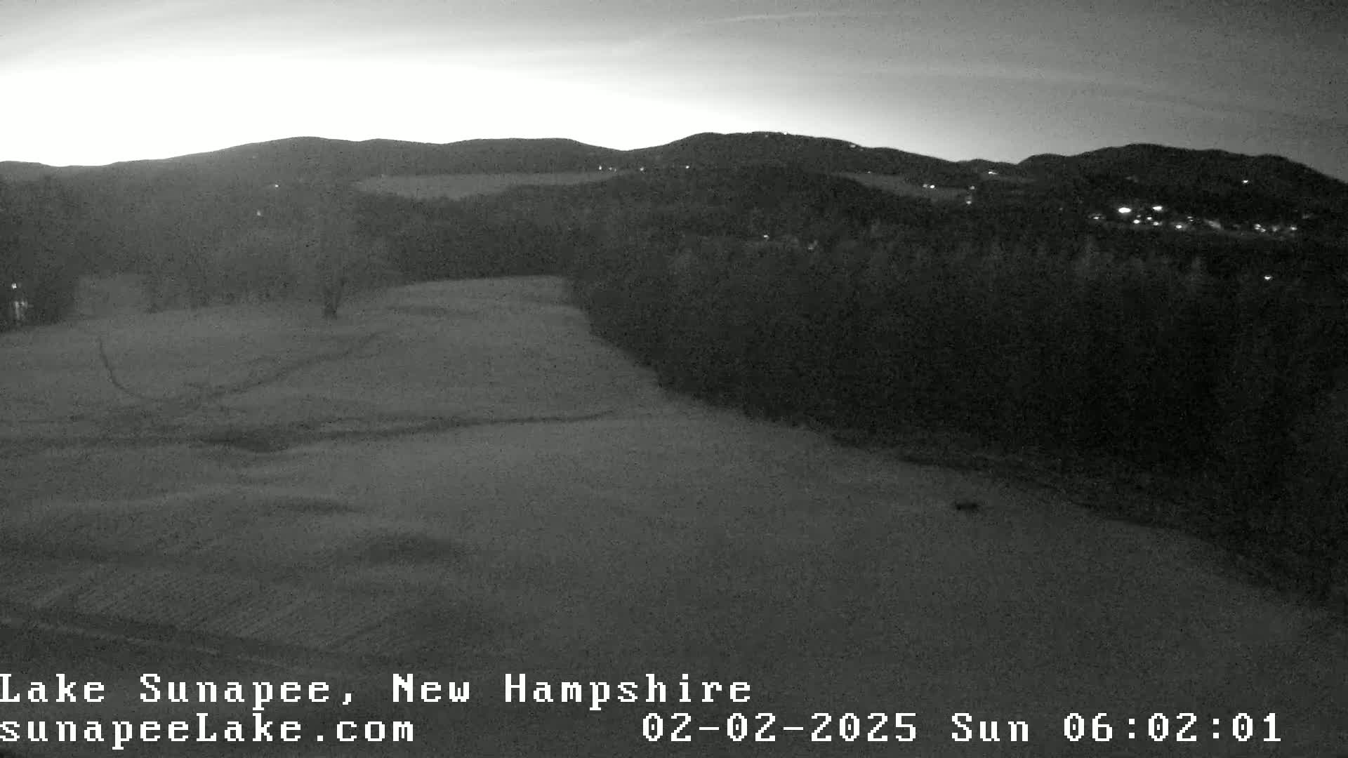 A nighttime, low-light grayscale image shows a wide, flat expanse of land, possibly a field or golf course, bordered by dark trees and distant hills under a dusky sky.