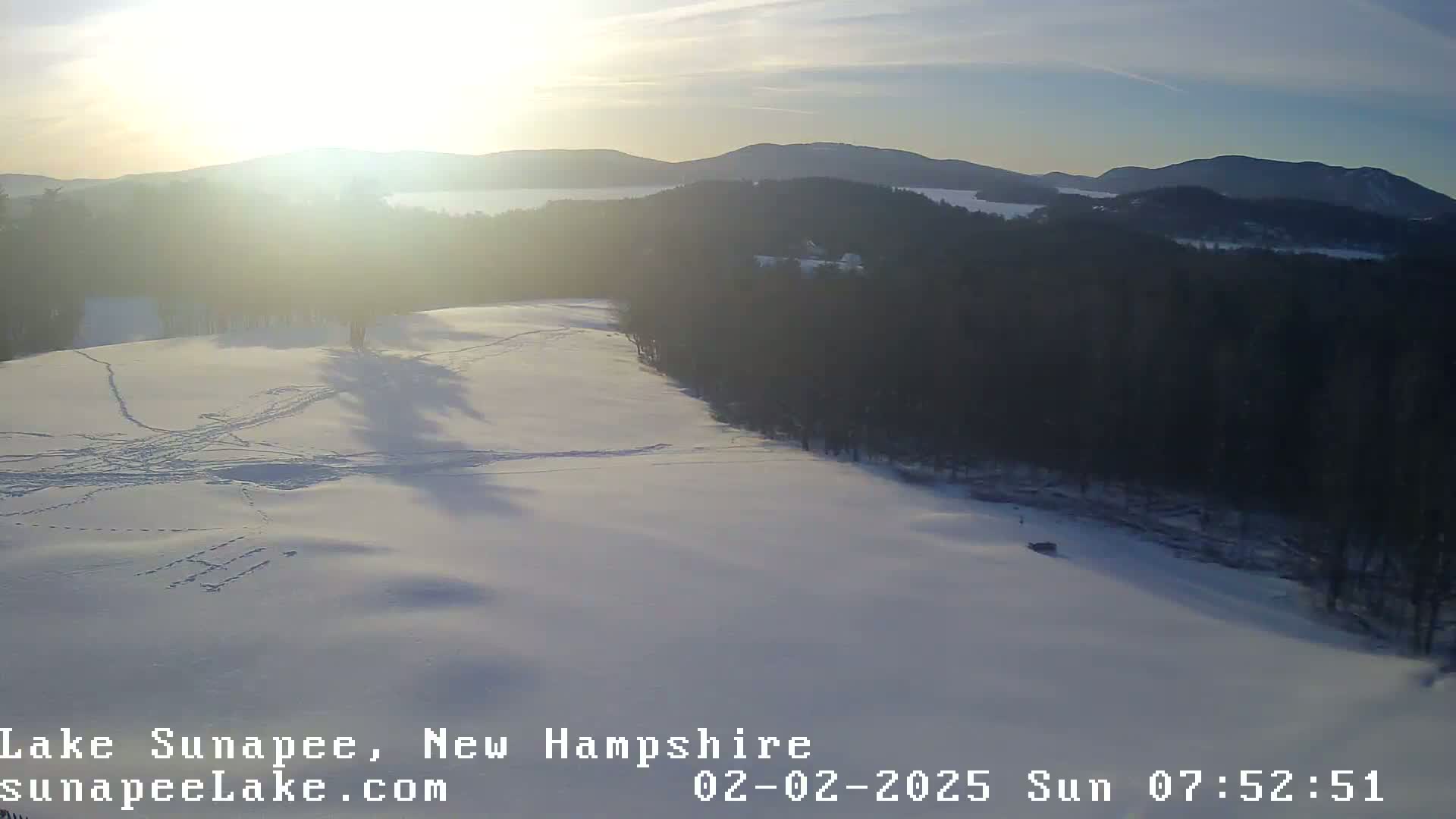 A snow-covered landscape with a bright sun shining over dark evergreen trees and distant hills.