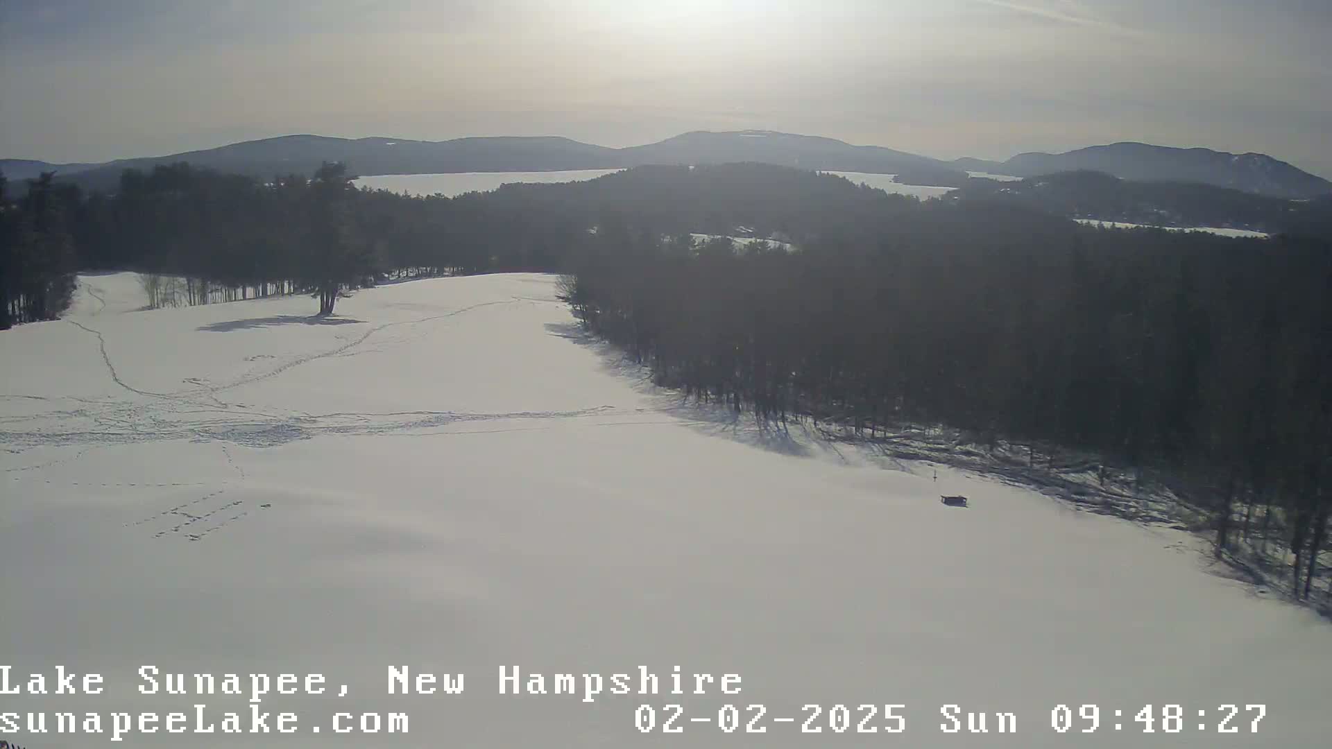 A snow-covered landscape with a line of trees and a distant lake is visible under a bright, sunny sky.