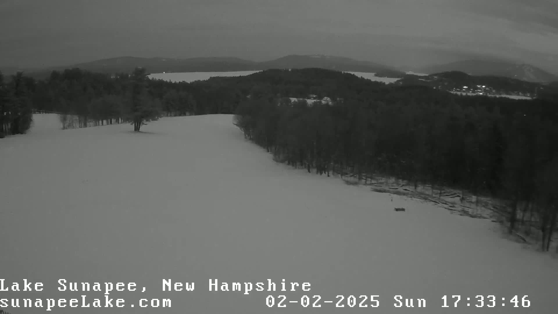 A snow-covered hillside descends toward a dark, tree-lined lake under a dim, overcast sky.