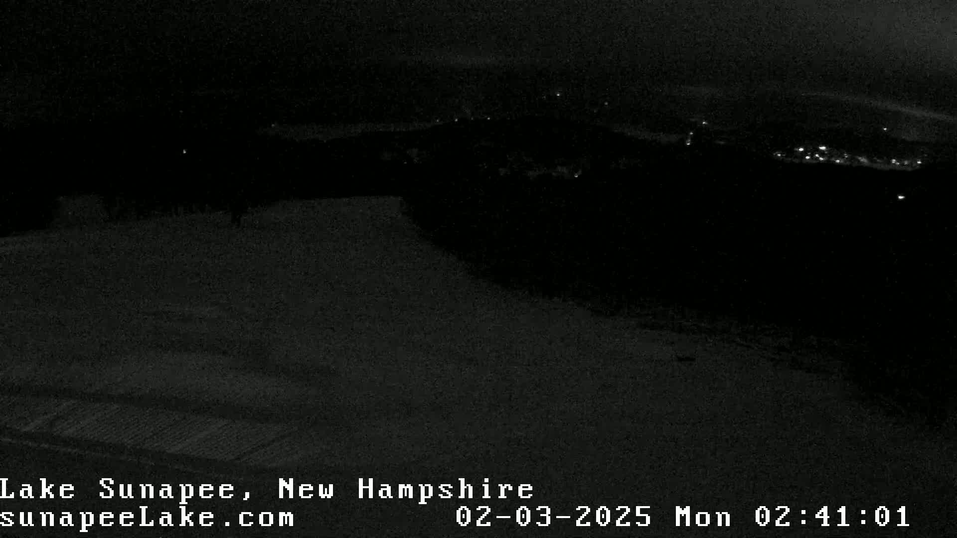 A dark, nighttime view of a snow-covered landscape with faintly visible lights in the distance.