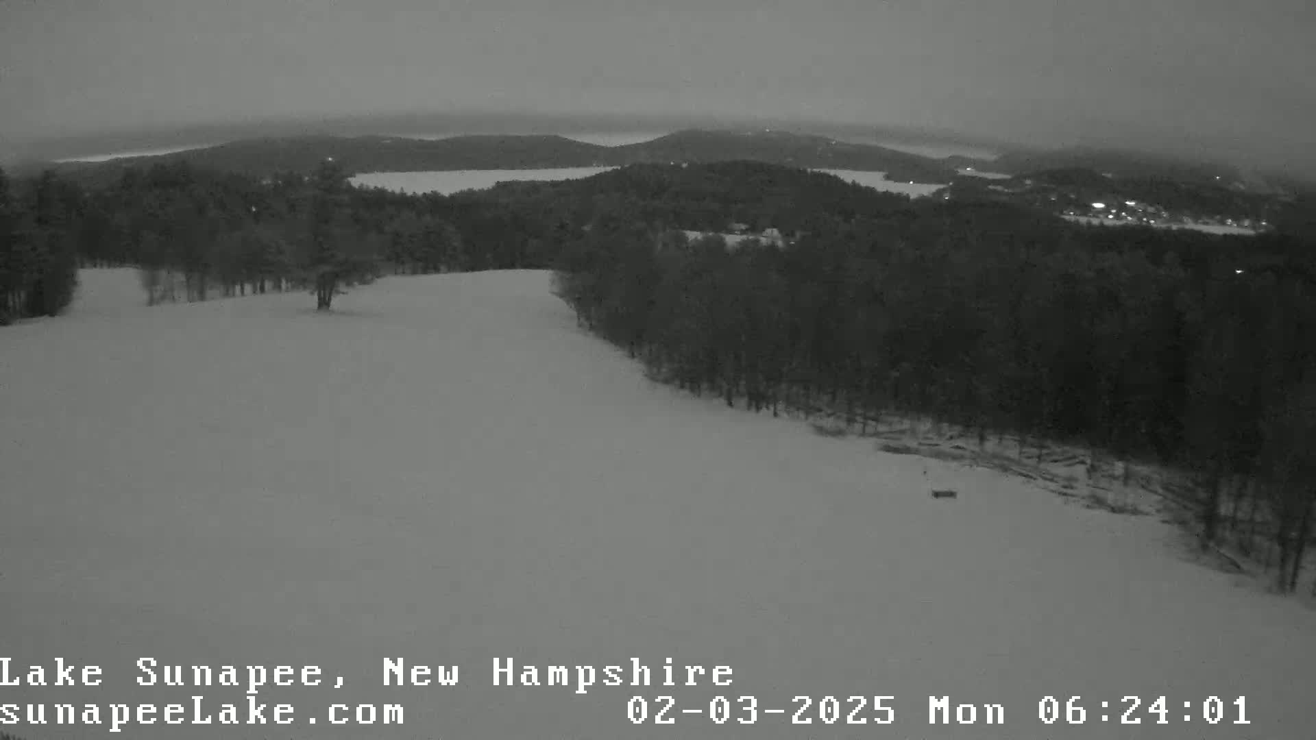 A snow-covered hill slopes down toward a dark, tree-covered landscape and a distant lake under a dim, overcast sky.