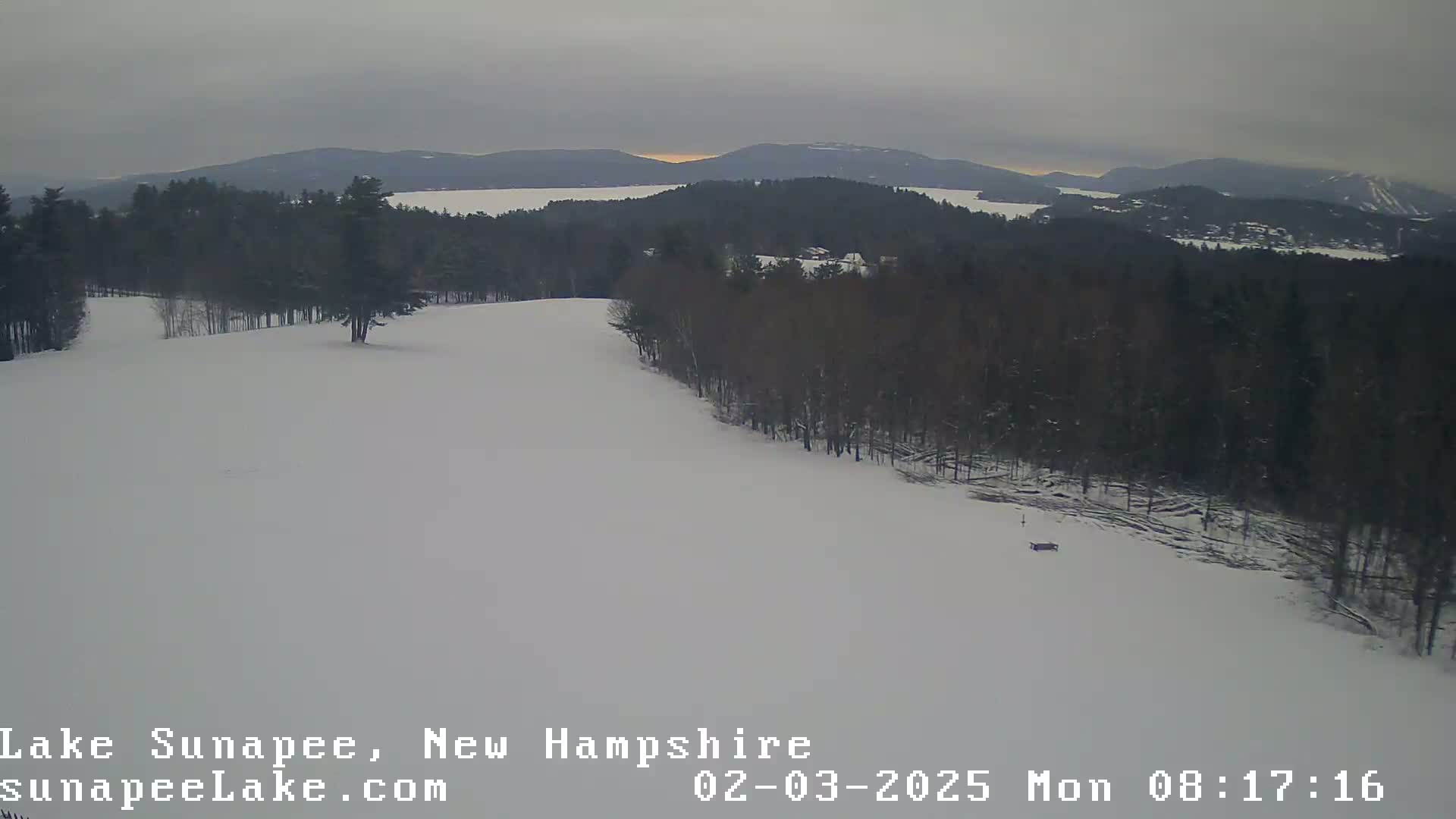 A snow-covered landscape shows a mostly-clear view of a lake and distant mountains under an overcast sky.