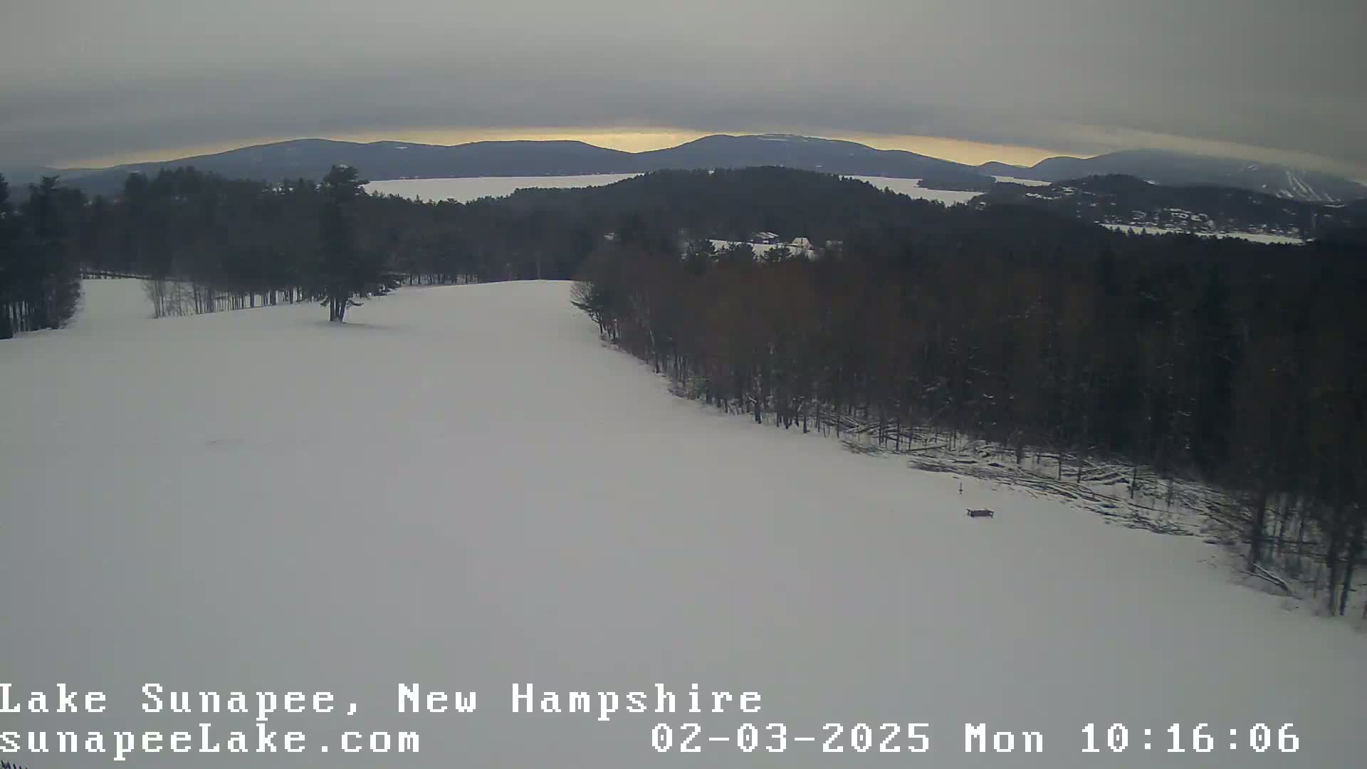 A snowy landscape shows a mostly overcast sky, a snow-covered hill, and a dark forest leading to a distant lake and mountains.