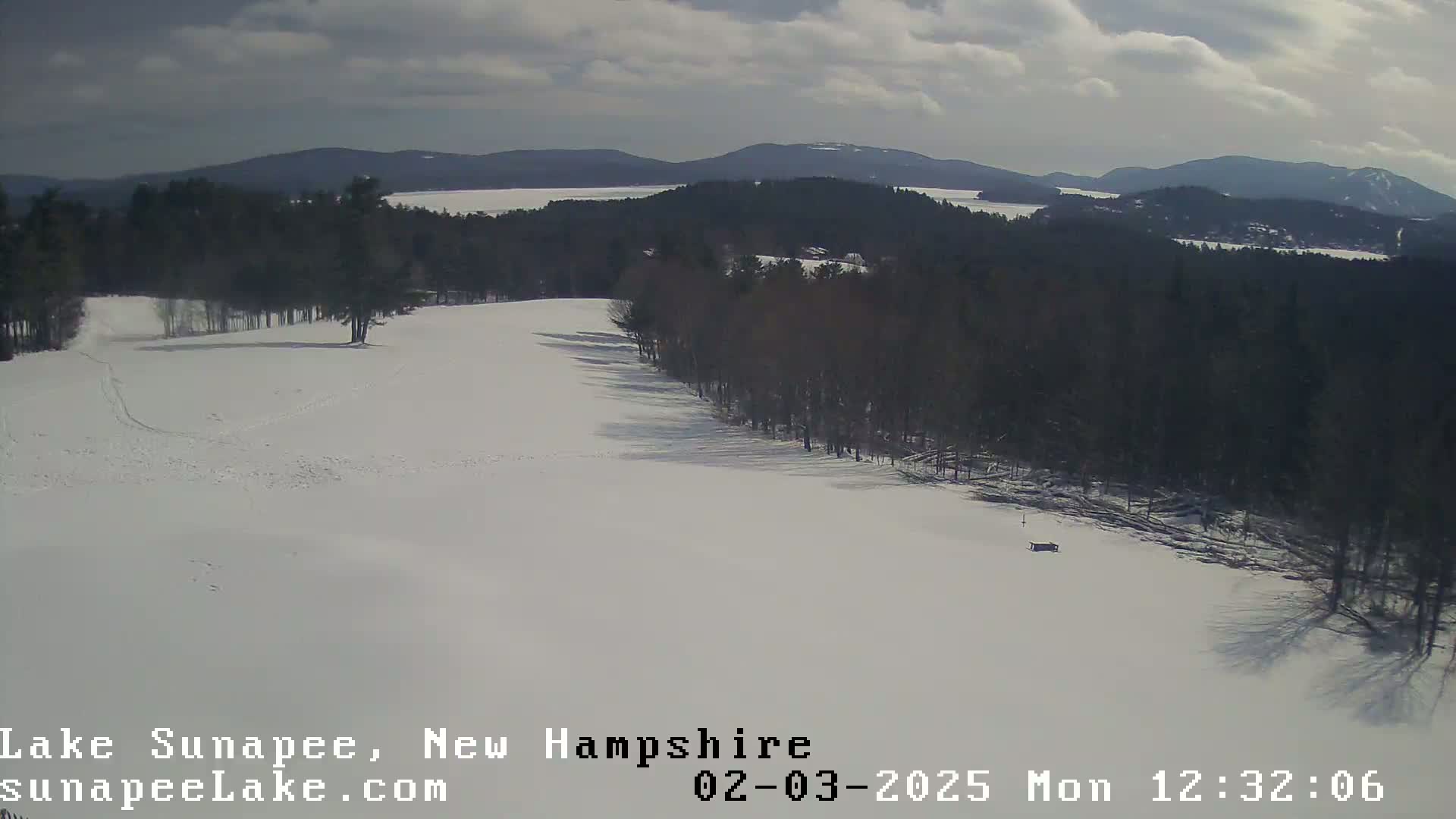 A snow-covered landscape with a line of trees, a distant lake, and mountains under a partly cloudy sky.