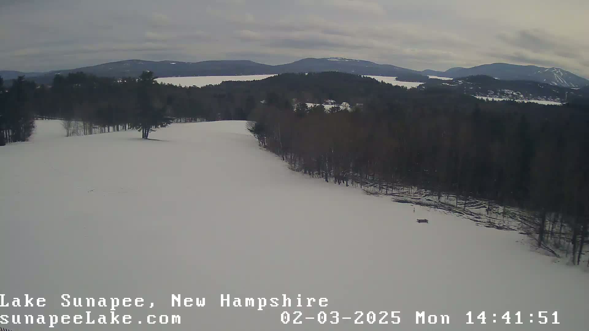 A snow-covered hill overlooks a lake and distant mountains under a cloudy sky.