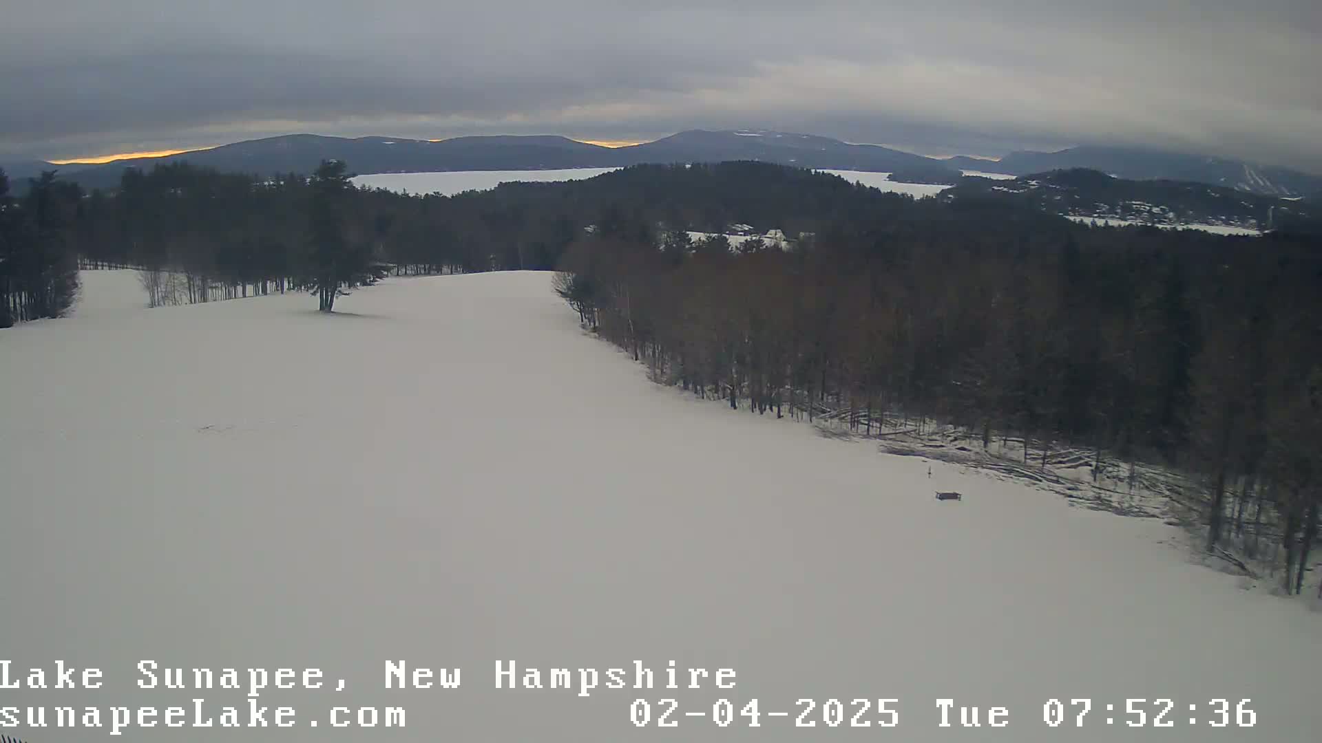 A snow-covered landscape shows a line of bare trees bordering a wide, open, snowy area that slopes gently downward toward a distant lake and mountain range under an overcast sky.