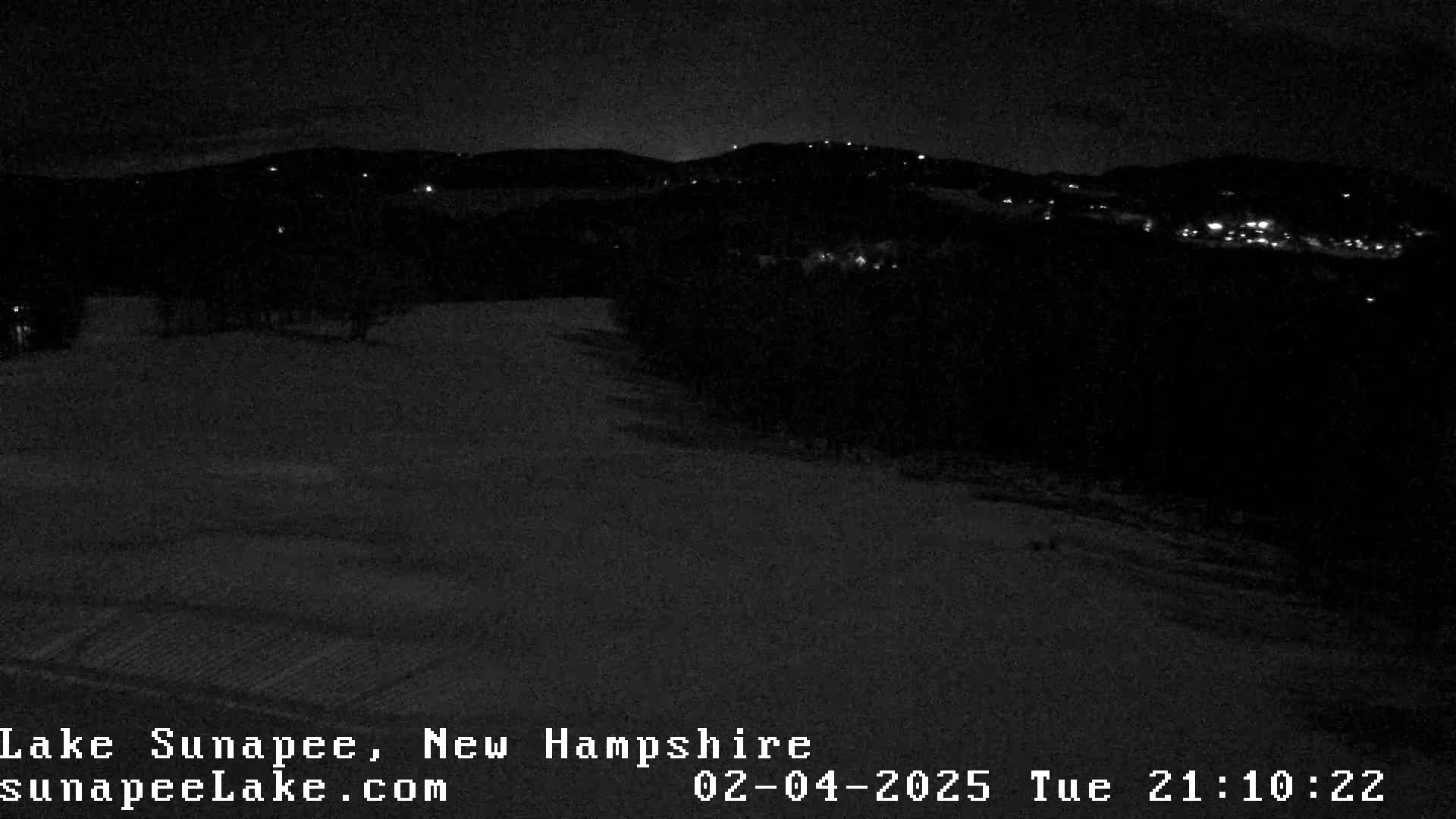 A nighttime, snow-covered landscape shows dimly lit buildings on a distant hillside under a dark sky.