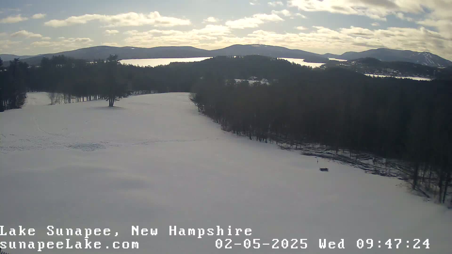 A snow-covered landscape with a partially frozen lake and dark evergreen trees in the background under a partly cloudy sky.