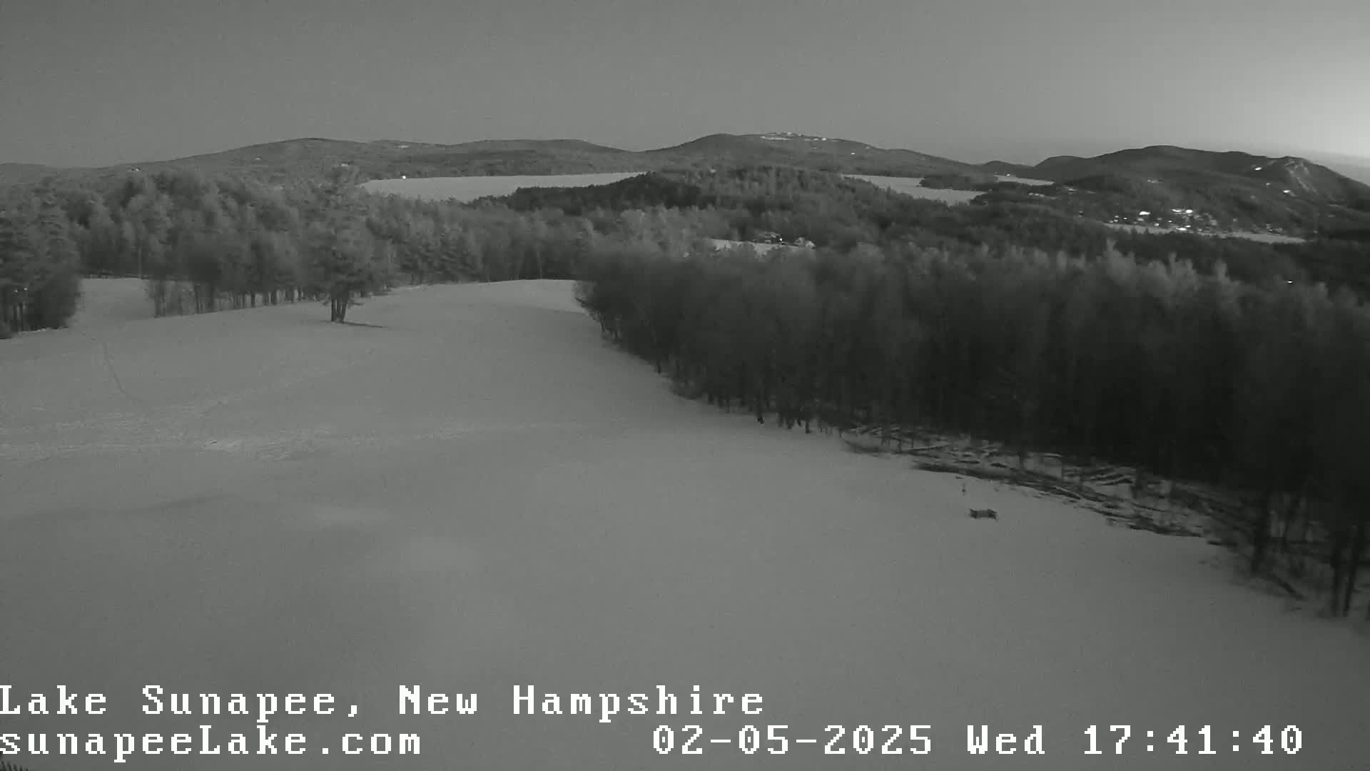 A snow-covered hillside with a line of trees at the bottom, overlooking a lake and distant mountains under a twilight sky.