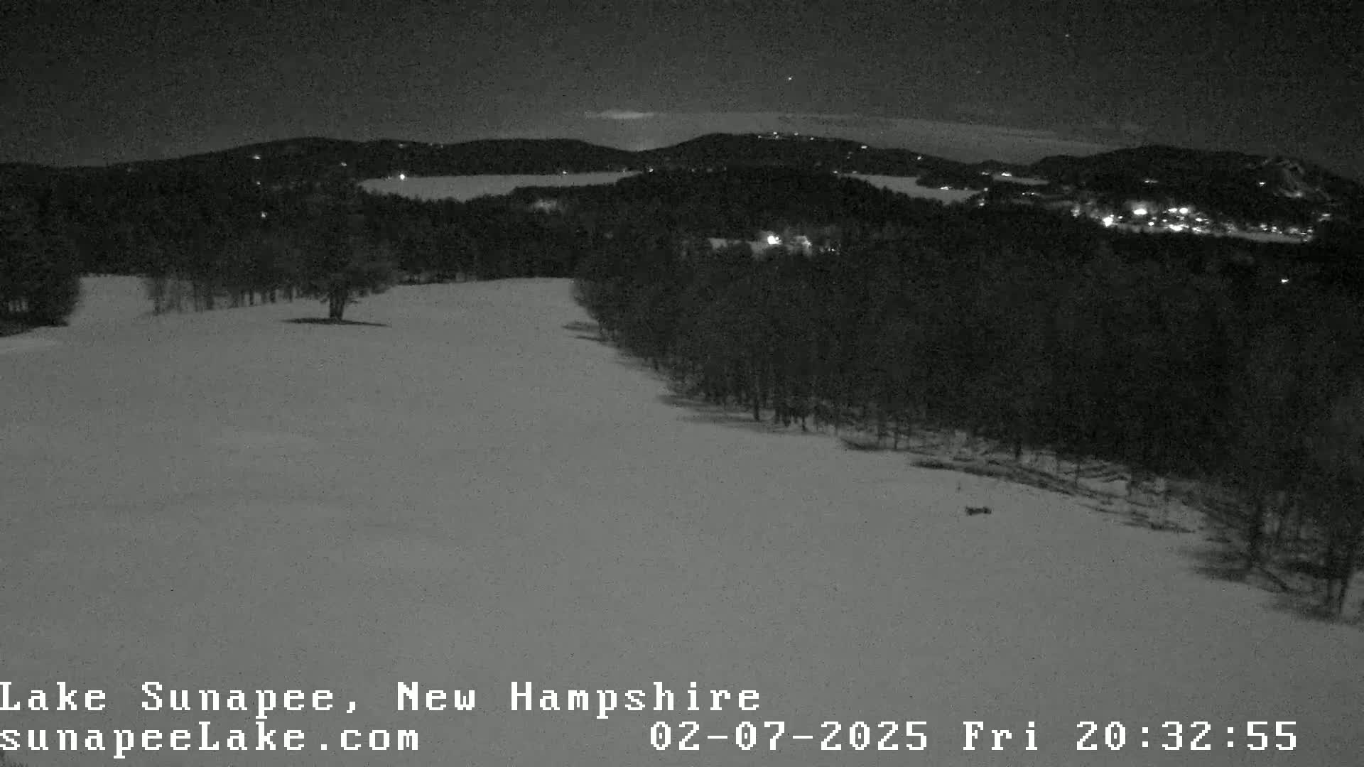 A nighttime, snowy landscape shows a hill covered in snow with a dark forest in the background, illuminated by faint lights.