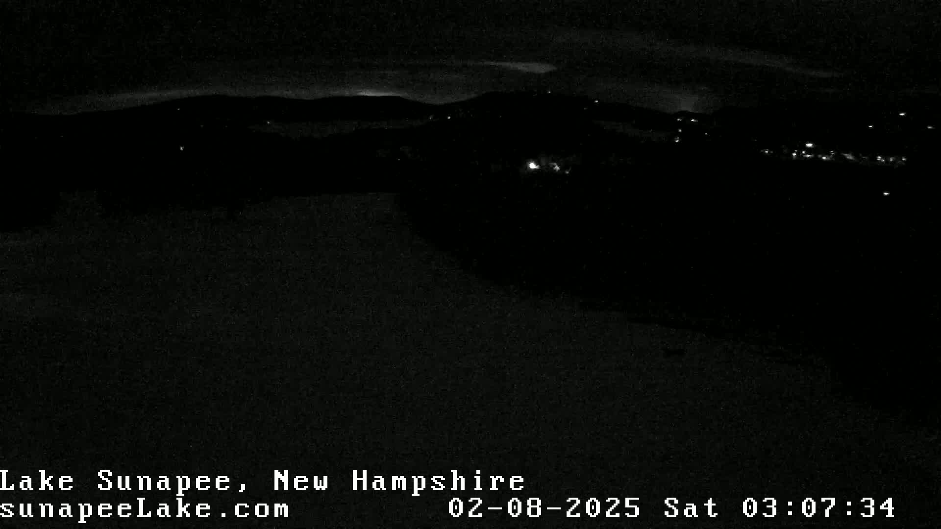 A nighttime long shot of a dark landscape with sparsely scattered lights in the distance under a mostly clear sky.