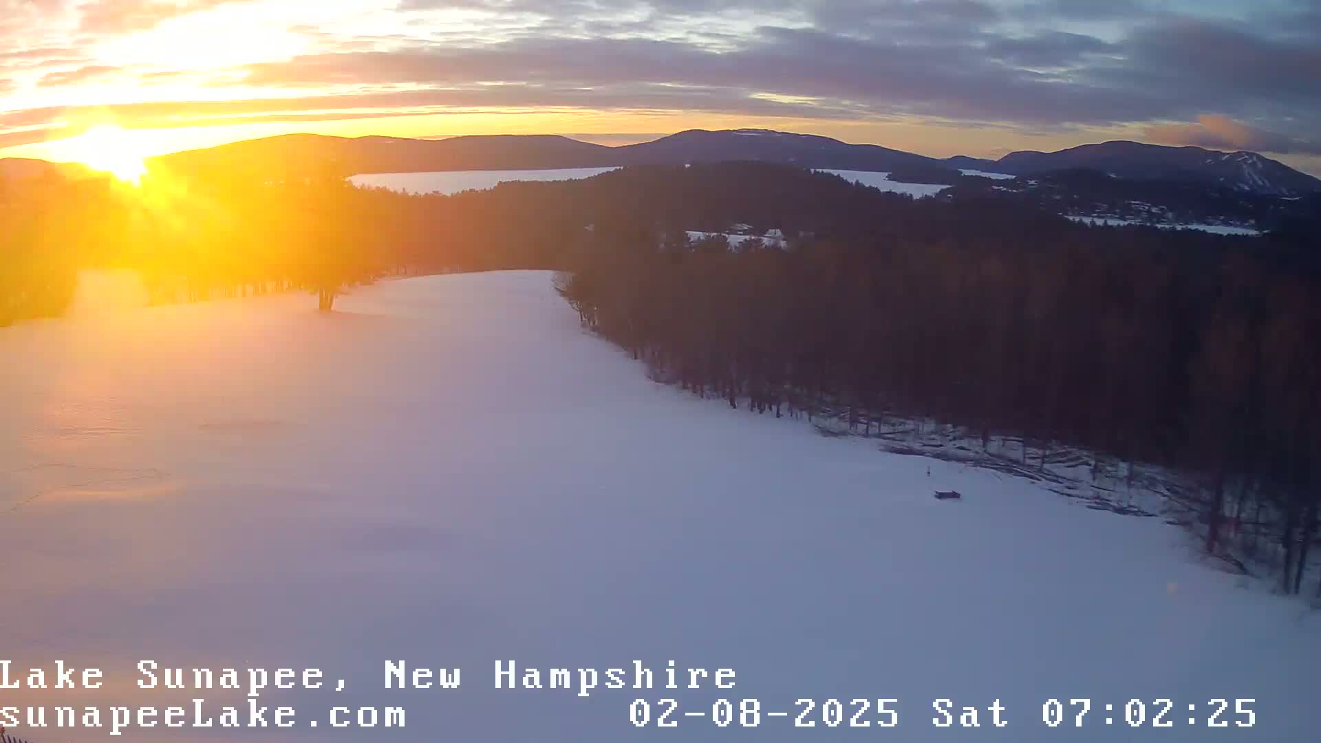 A snowy landscape at sunrise, featuring a lake and forested hills in the background.