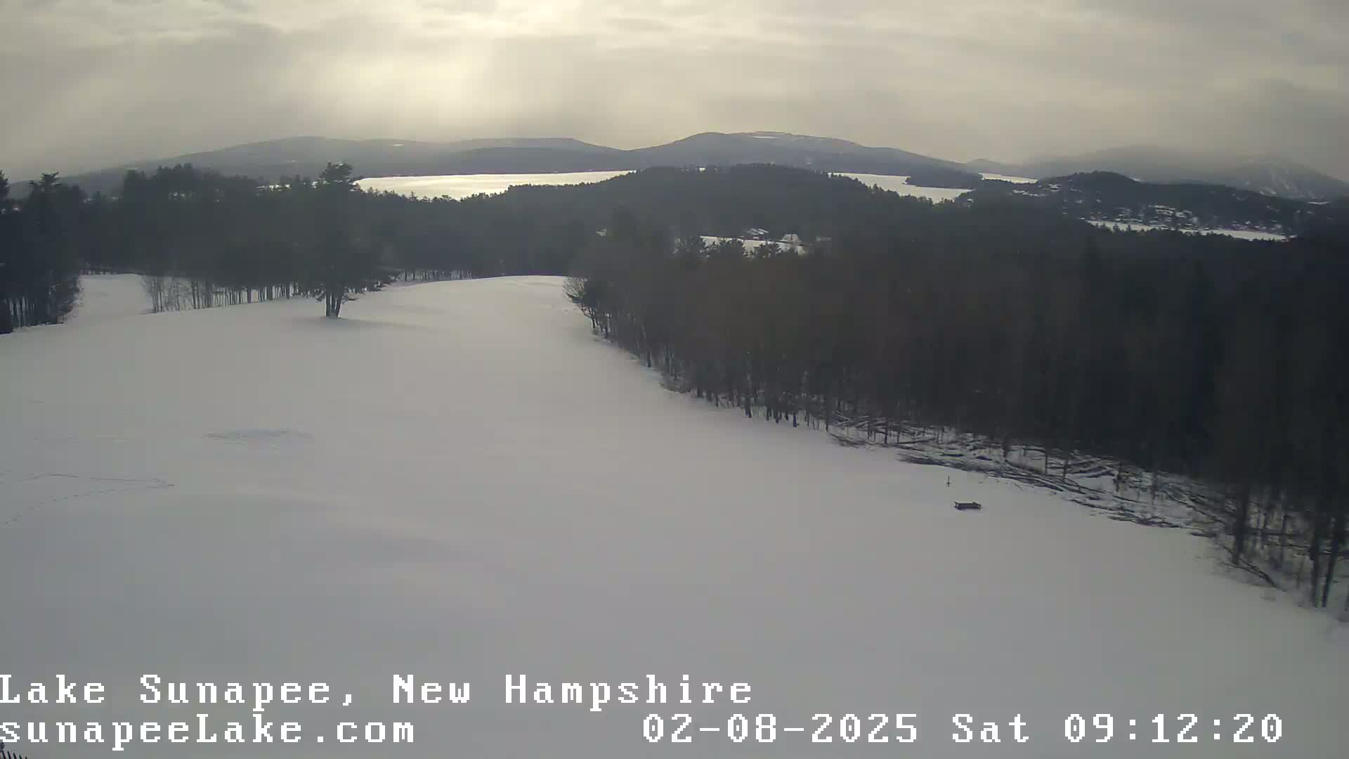 A snow-covered landscape shows a gently sloping hill leading to a lake and distant mountains under an overcast sky.