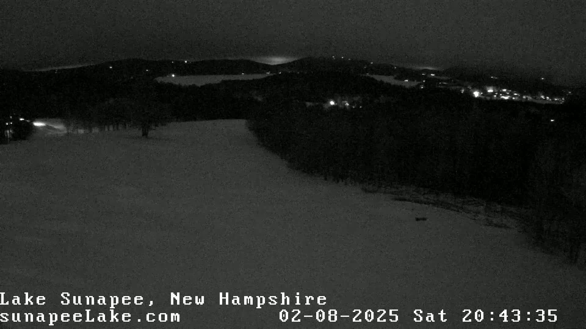 A nighttime, snowy landscape shows a hilly area with scattered lights from a town in the distance.