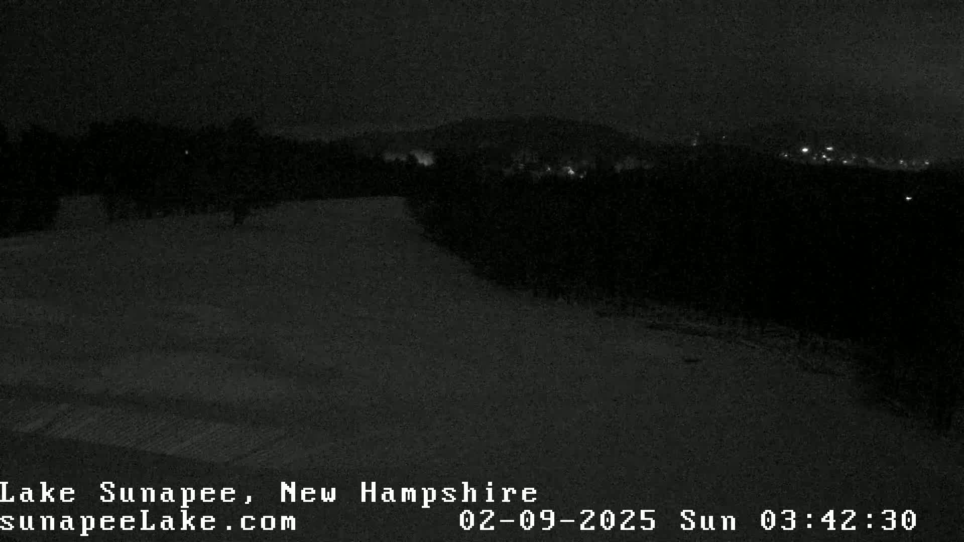 A nighttime, snowy landscape shows a dark, snow-covered ground leading towards a distant, dimly lit town nestled among dark trees under a dark sky.