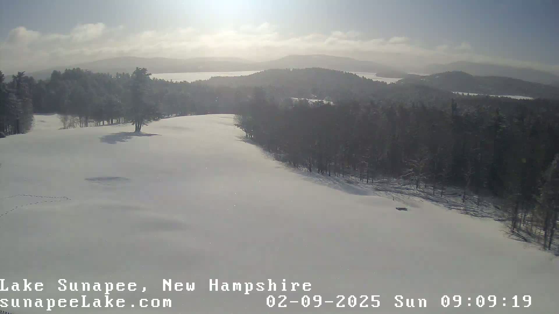 A snow-covered landscape, featuring a hilly terrain with a forest of dark trees in the background, is visible under a mostly sunny sky with some clouds.