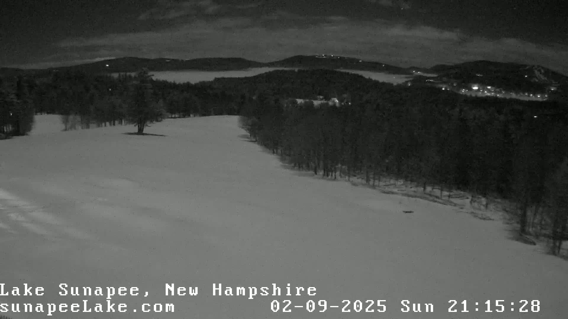A snow-covered hill at night, bordered by dark trees, with a hazy, low-lying cloud or fog obscuring the distant mountains.