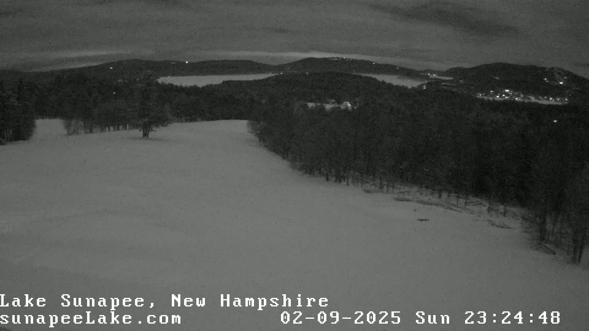 A snow-covered hill and forest overlook a dark, distant lake under a night sky.