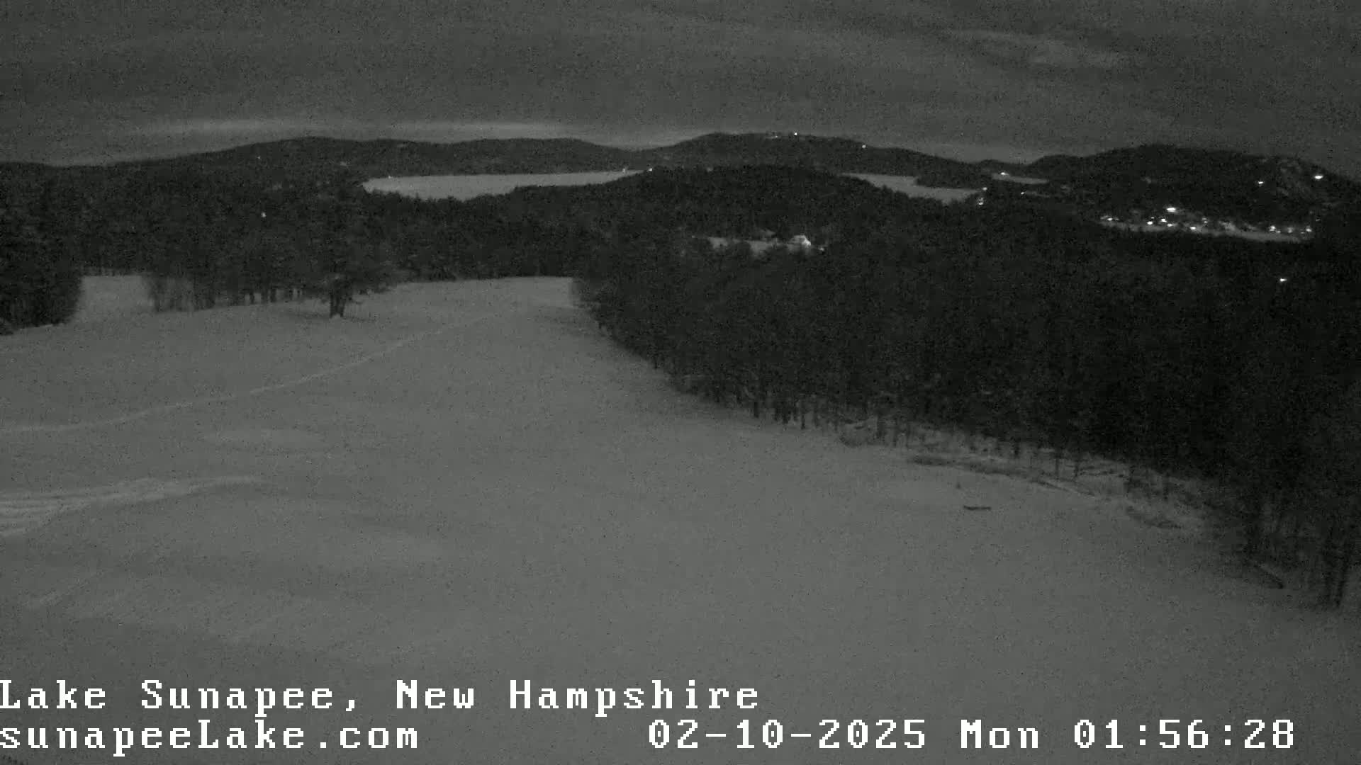 A snow-covered landscape at night, featuring a dark forest and a body of water in the distance under a cloudy sky.