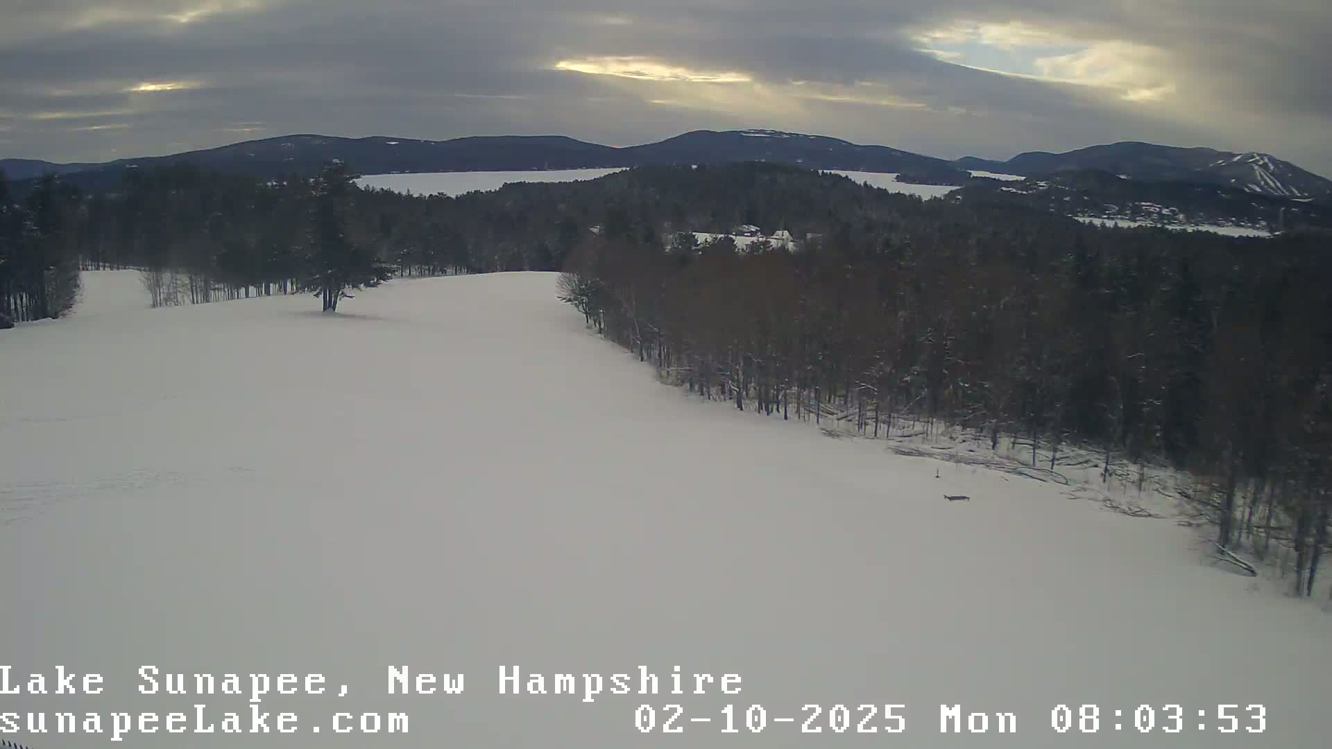 A snow-covered landscape shows a mostly cloudy sky over a lake and distant mountains, with a line of trees separating a snowy field from the water.