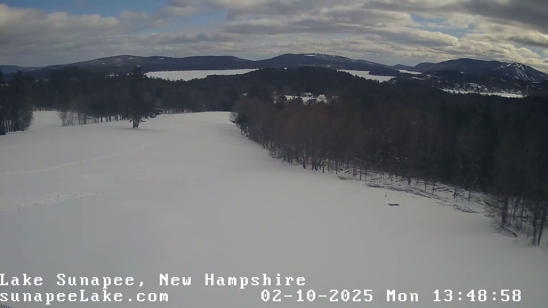 A snow-covered landscape with dark, leafless trees and a body of water visible in the distance under a cloudy sky.