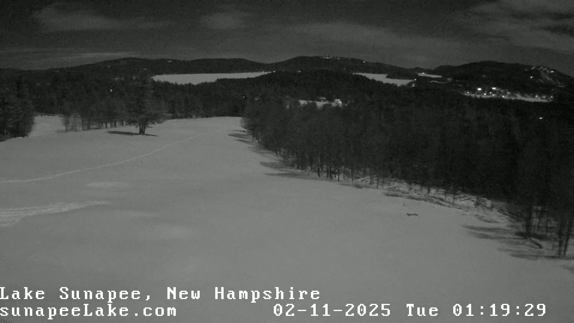 A nighttime, snow-covered landscape shows a dark forest and a body of water in the distance under a cloudy sky.