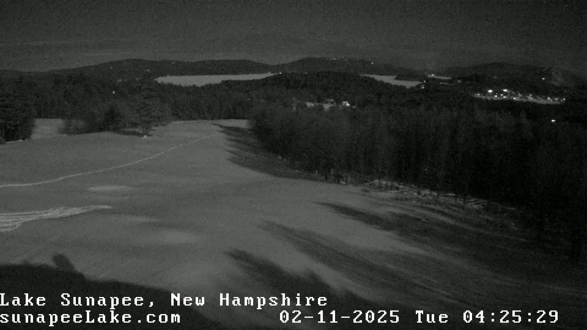 A nighttime, snow-covered hill slopes down toward a dark, wooded area with a body of water visible in the distance under a clear, dark sky.