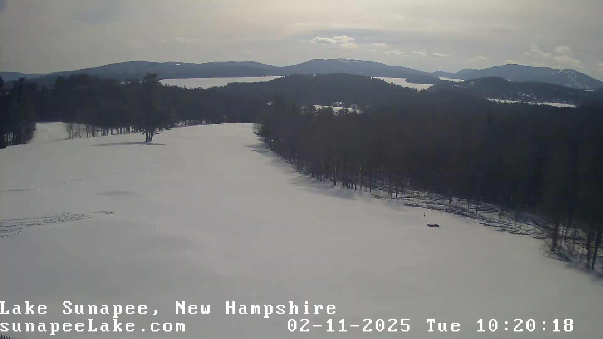 A snow-covered landscape with a line of trees bordering a body of water in the background under a partly cloudy sky.
