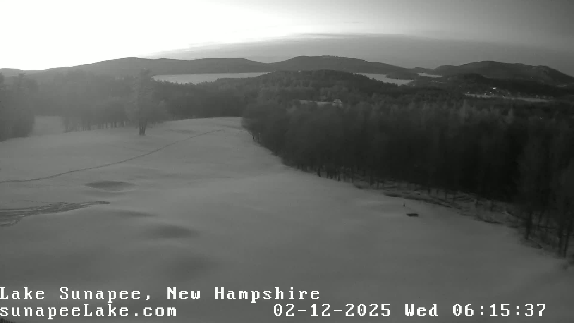 A snow-covered landscape, showing a hilly terrain with a line of trees in the background, under a twilight sky.