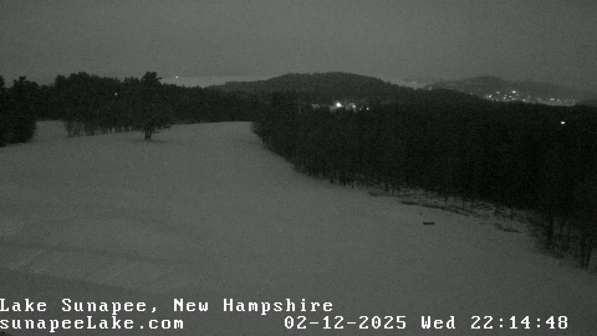 A snow-covered landscape at night, with dark trees and distant lights visible under a dark sky.