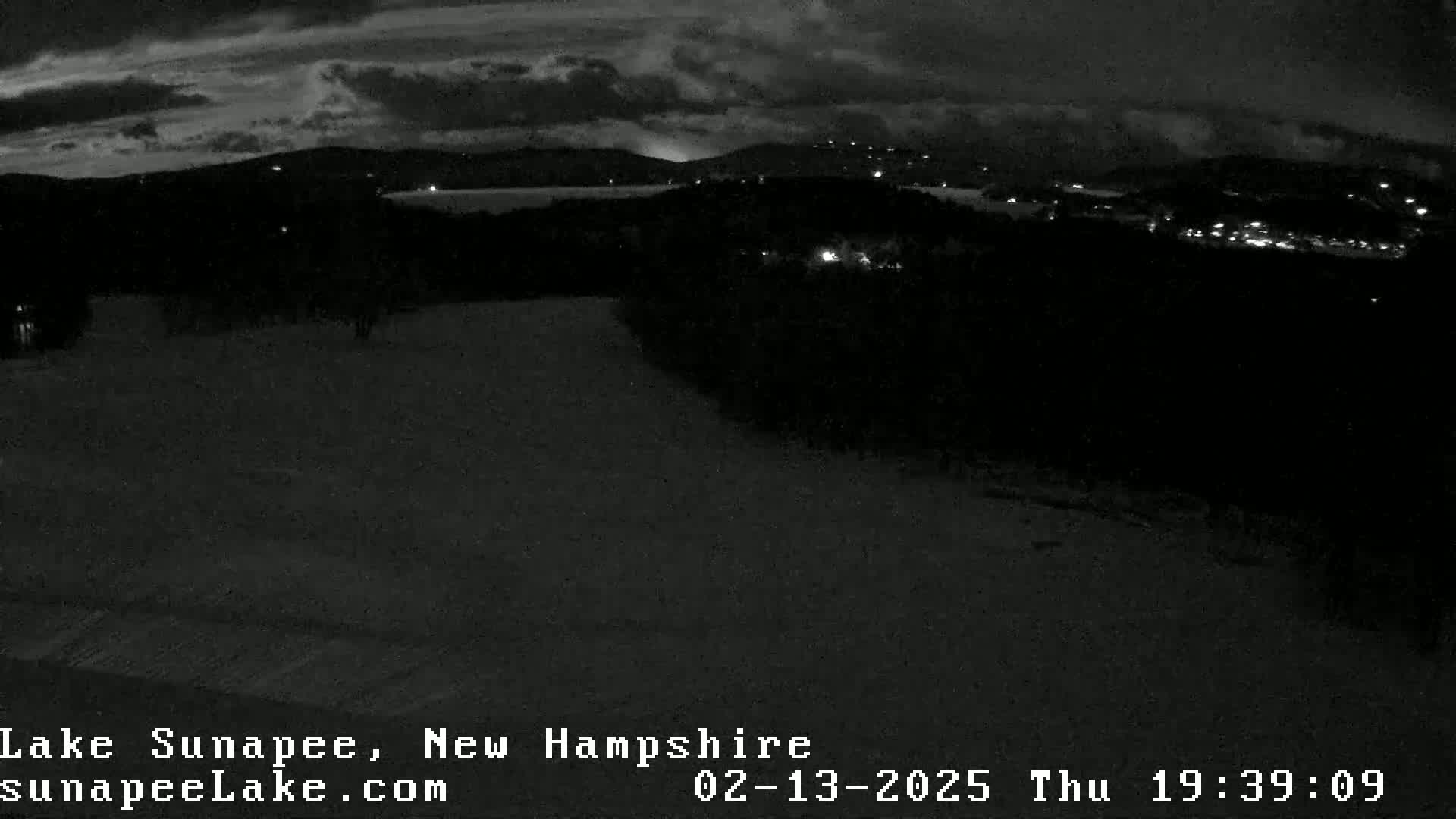 A nighttime, snowy landscape overlooking a dark lake and distant, faintly lit town under a cloudy sky.