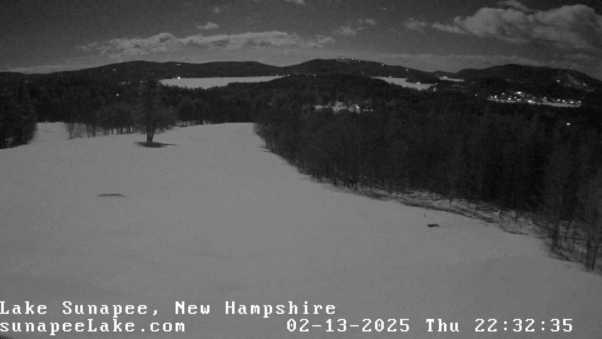 A snow-covered landscape at night, under a partly cloudy sky, shows a dark, wooded area with a body of water visible in the distance.