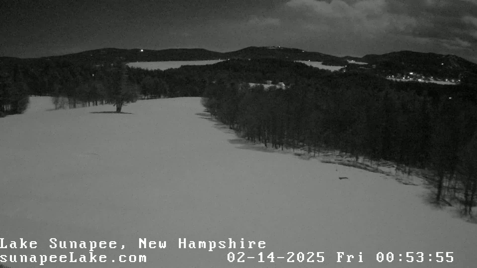 A snow-covered landscape at night shows a dark, wooded area and a body of water in the distance under a cloudy sky.