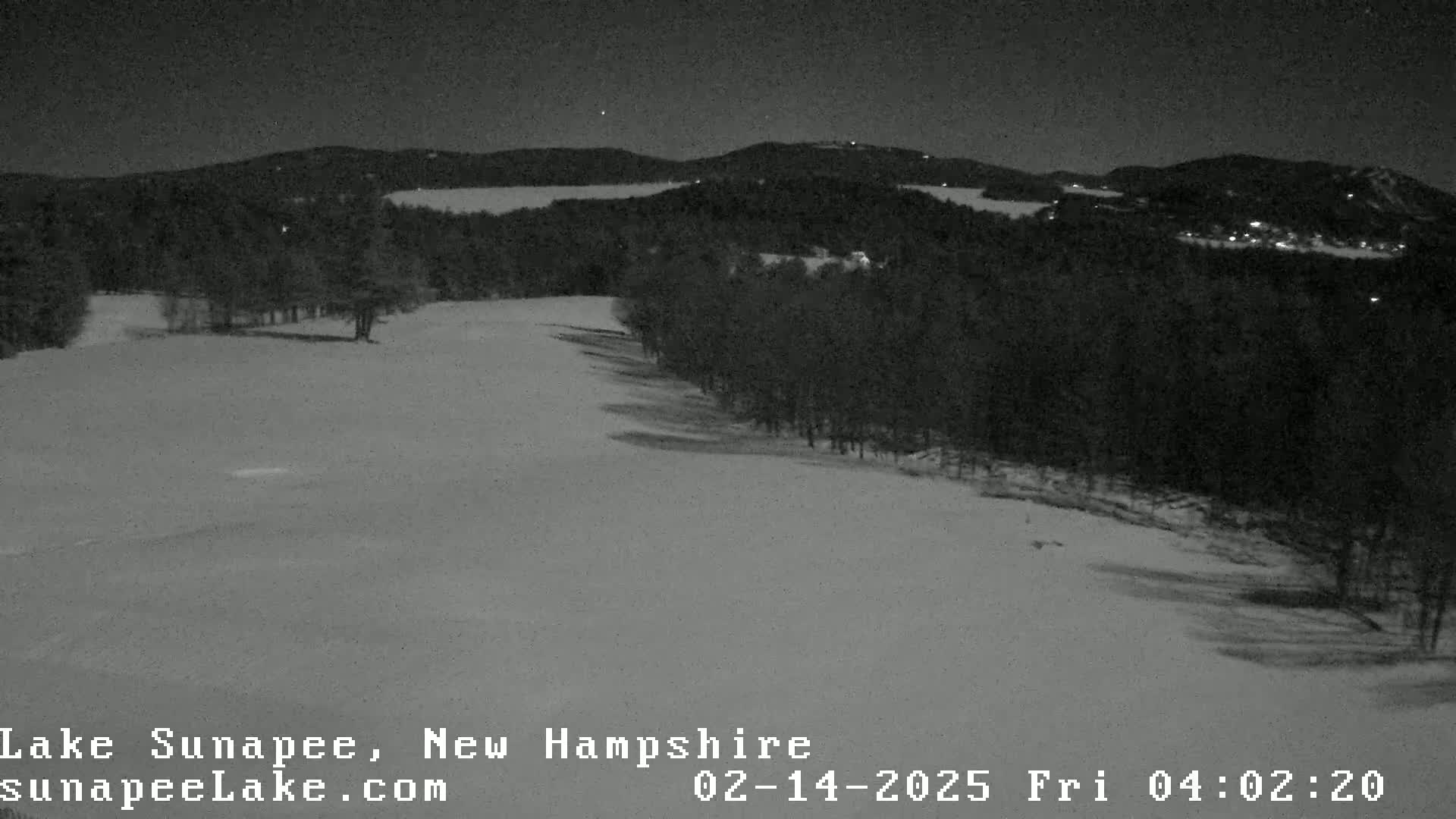 A snow-covered landscape at night, showing a line of trees bordering a wide, flat expanse of snow under a dark sky.