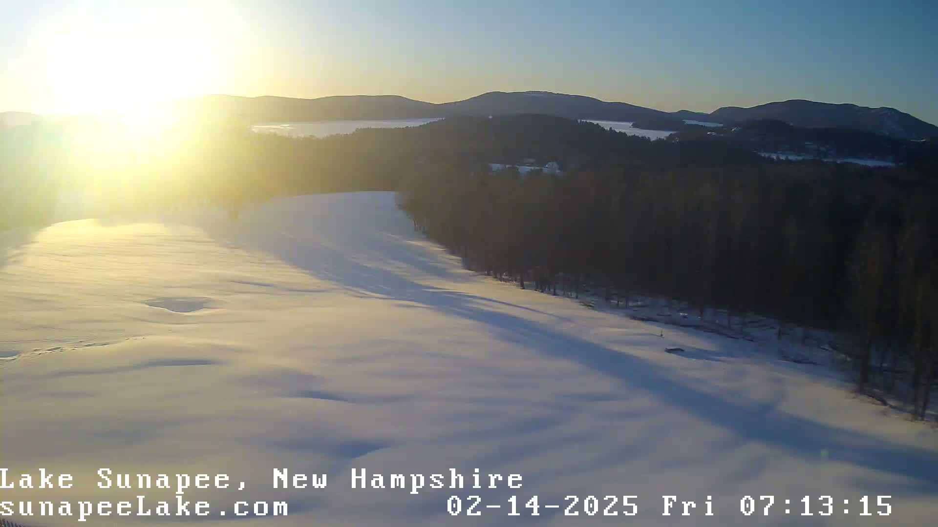A snow-covered landscape with a sunlit, slightly hazy sky in the background, overlooking a dark forest and a partially visible lake.