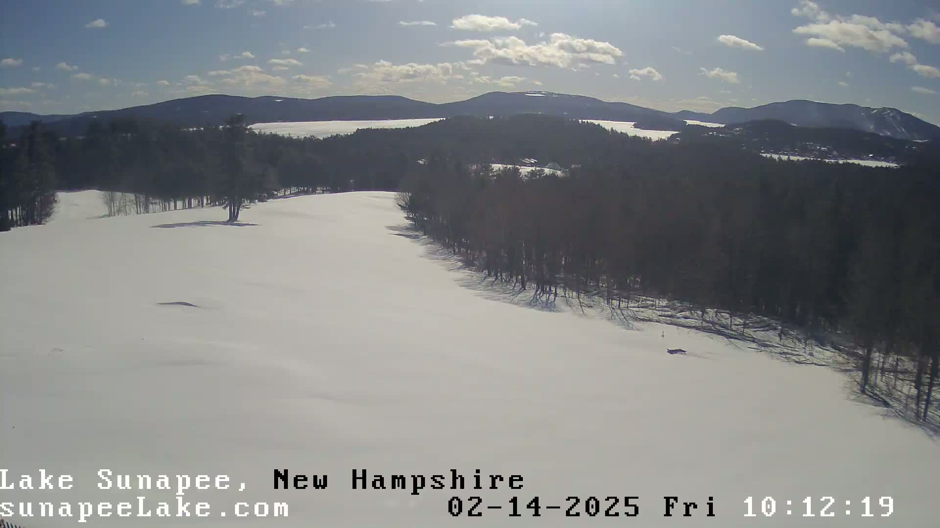 A snow-covered landscape with a line of dark trees bordering a mostly flat, snow-covered field under a partly sunny sky, overlooks a distant lake and mountains.